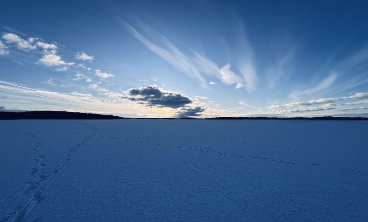 A frozen lake under a winter sky, with ski tracks and footprints leading across the snow toward the far shore.